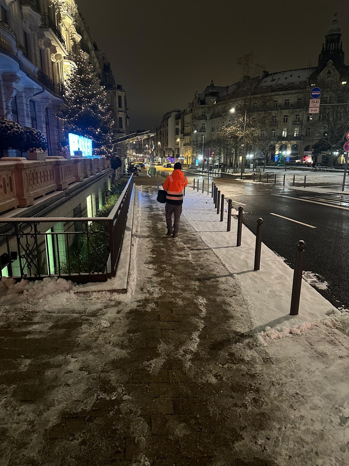 Mitarbeiter von Eymen Galabau streut in Warnkleidung bei Nacht einen geräumten Gehweg in der Innenstadt